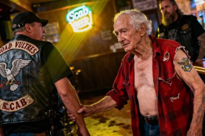 Beneath the jaundiced glow of a seaside tavern, where old ghosts and cheap beer mingle, an old man’s torn shirt revealed more than just a faded tattoo—it unearthed a covenant, reminding us that true legends don’t roar; they endure.